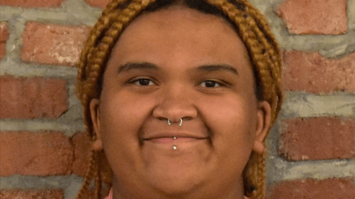 A person with braided hair wearing a pink blazer and a white top, smiling in front of a brick wall. They have a septum piercing and a labret piercing.